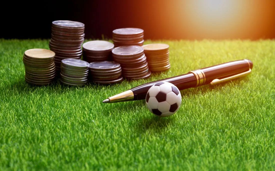 Geldmünzen, Füller und Fußball auf einem Rasen als Symbol für Lizenzen im Sportwetten Vergleich